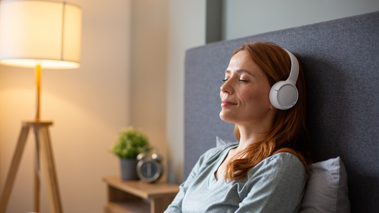 A woman lying back in bed with her eyes closed listening to audio from a pair of white headphones.