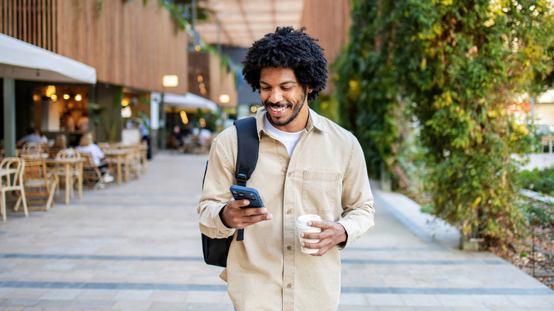 A man using his phone outdoors, while holding a coffee cup in the other hand.