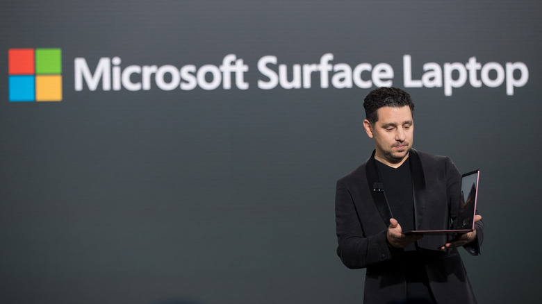 Presenter holding an opened Microsoft Surface Laptop in front of a Microsoft logo display.