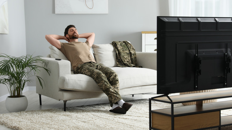 A man with his hands behind his head relaxing on a living room couch in front of a TV
