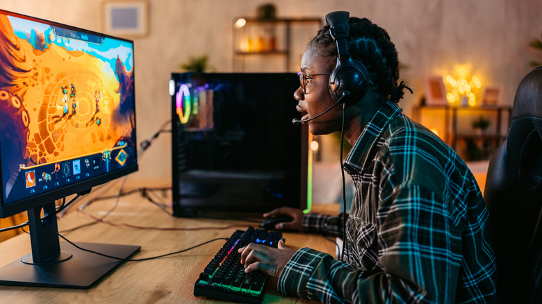 A person sitting at a desk, looking at a monitor while playing game with keyboard and mouse
