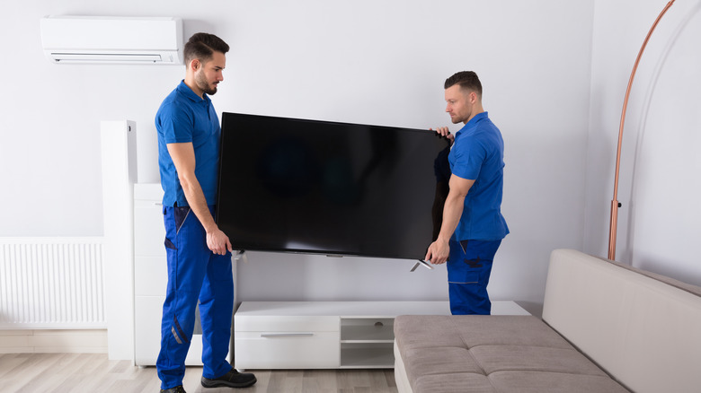 Two workmen in blue attire moving a TV in a room.