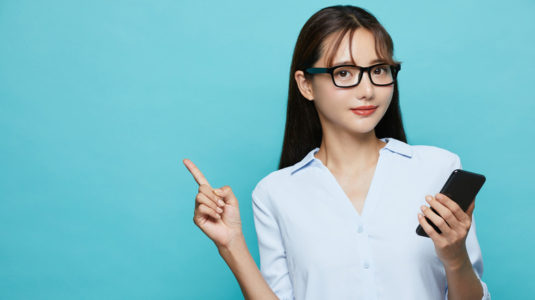 Woman holding smartphone and point upward with opposite hand on teal background