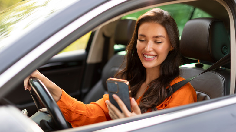 A person with a smile on their face looking directly at their cellphone as they sit behind the wheel of an automobile
