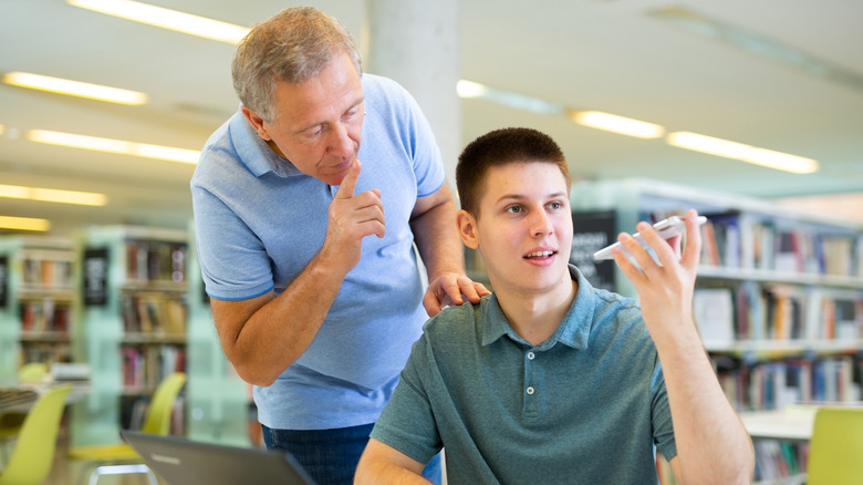 An individual using a speakerphone in a public library, holding the cellphone in a hand while another patron shushes the cell user.