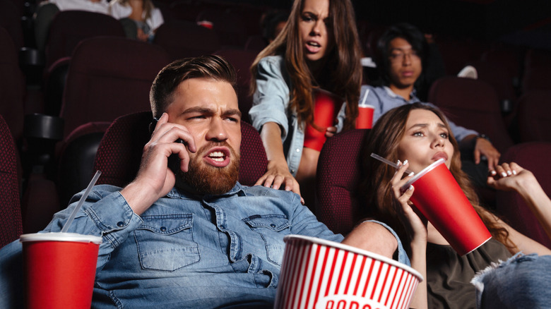 An individual in a blue shirt is on their cellphone in a movie theater, while a person in the row behind them is attempting to get their attention by placing their right hand on the cellphone user's shoulder