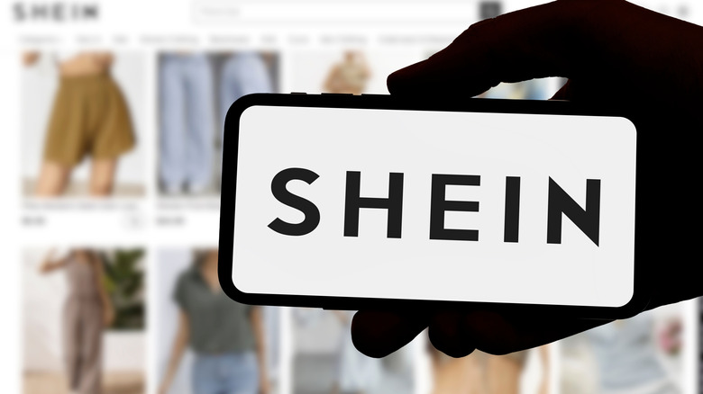 A phone displaying the word Shein being held in front of a screen showing Shein's web page