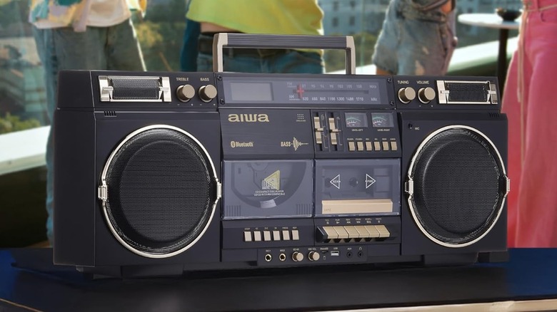 A black Aiwa Retro Boombox with gold trim sits at the forefront of a party that's occurring in the background