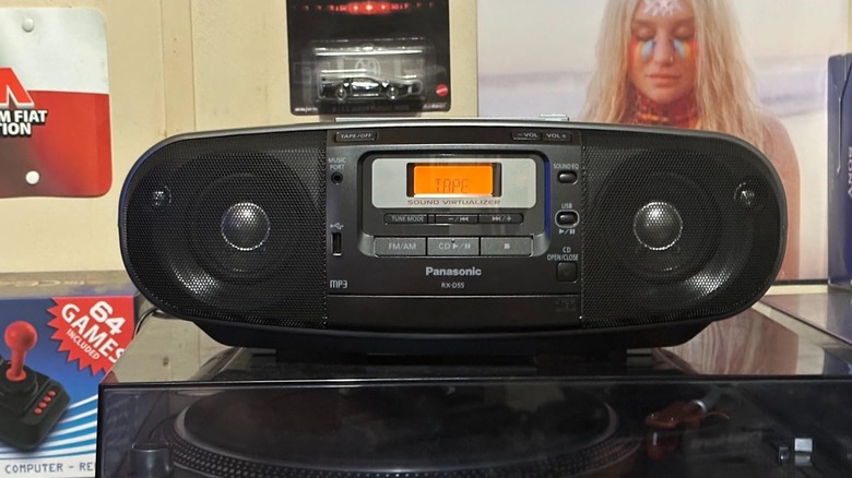 A Panasonic RX-D55GC-K Boombox sits on top of a record player, the background has a poster and a small model car in its packaging attached to the wall