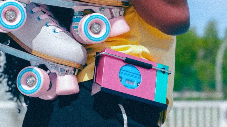 The We Are Rewind Portable Cassette Player in neon pink and blue hangs from an individual's waist as they carry a pair of colorful rollerskates