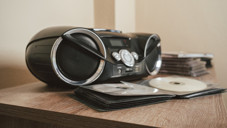 A black CD player boombox sits on a table next to a pile of CDs and a CD booklet holding two discs