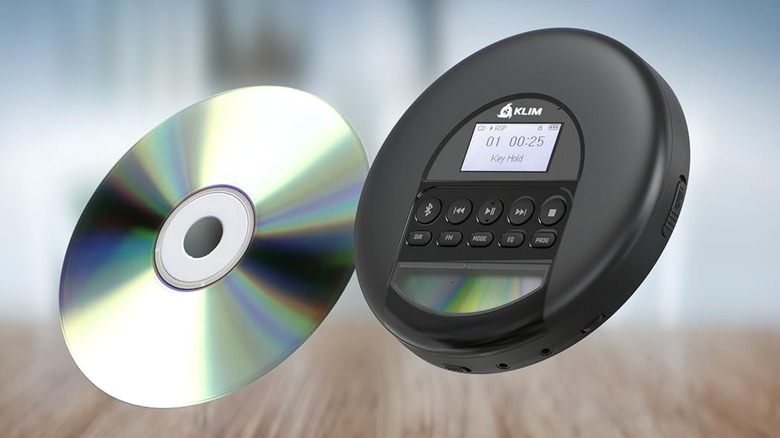 A black Klim Nomad CD player is superimposed over a blurry image of what appears to be a room, there is also a CD next to the player