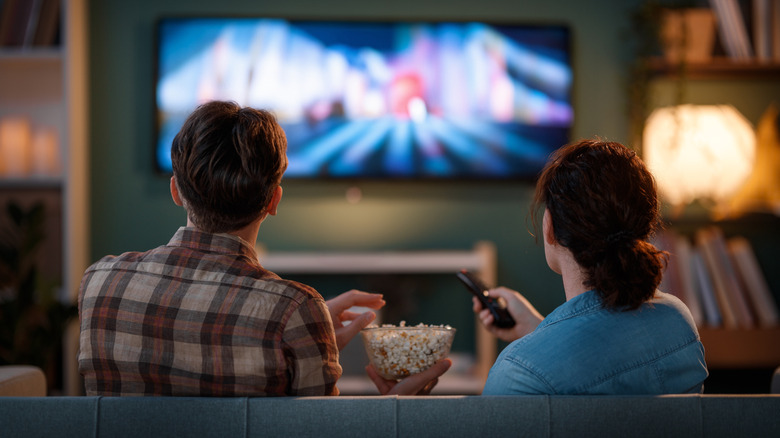 Couple watching TV together on a couch, holding popcorn and a remote.