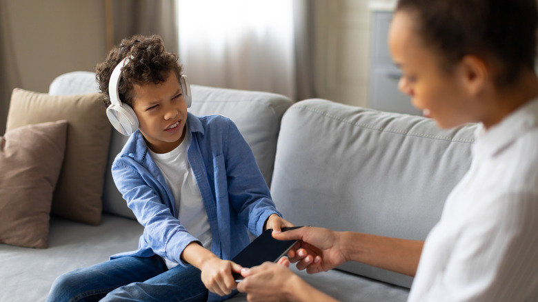 curly-haired child wearing headphones looks at his mother angrily as she takes his tablet away from him
