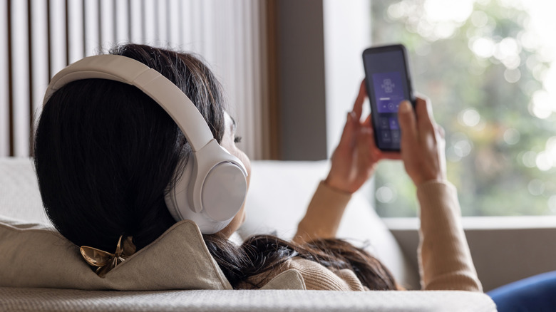 A behind-the-shoulder view of an individual sitting on a couch as they wear a pair of white headphones, the person is holding a cellphone with both of their hands