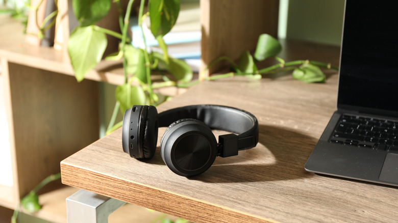 A pair of black wireless headphones sit next to a laptop on a wooden surface, there is at least one potted plant in the photo