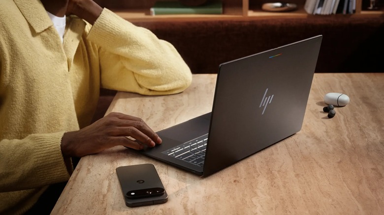 A user in a yellow sweater sits at a table in front of a black HP Googlebook 14, there is also a pair of Google earbuds outside of their white charging case alongside a black Google Pixel phone sitting on the table