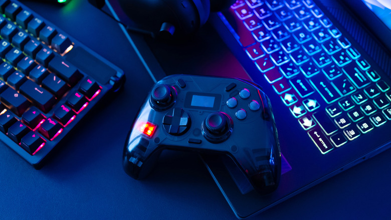 Game controller placed between an RGB keyboard and laptop on a gaming desk.