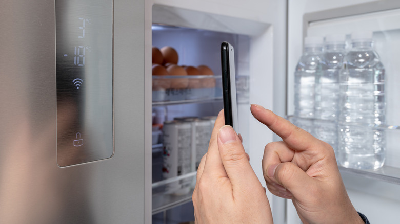 Person using a smartphone beside an open smart refrigerator with a Wi-Fi display panel.