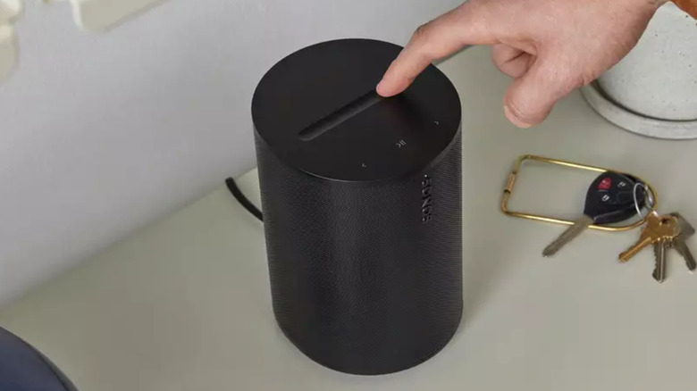 A black Sonos Era 100 SL Wireless Smart Speaker on a table being touched by a person's hand.