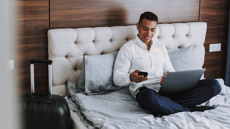 person sitting on a hotel bed with a laptop on his lap, a smartphone in hand, and luggage next to the bed