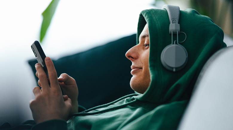 A person in a green hoodie wears a pair of light green headphones on their head as they keep both hands on a smartphone, notable, the person has their headphones over the hoodie on their head