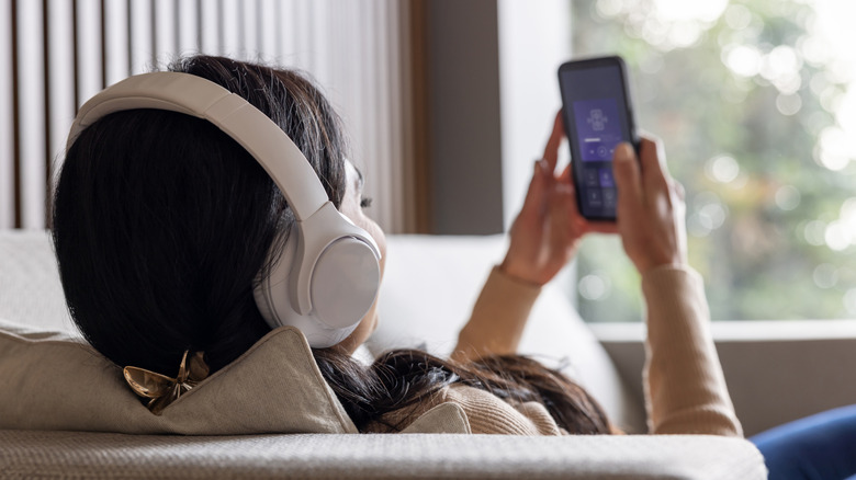 A behind-the-head shot of an individual wearing white headphones as they sit on a white couch and have both of their hands on a smartphone