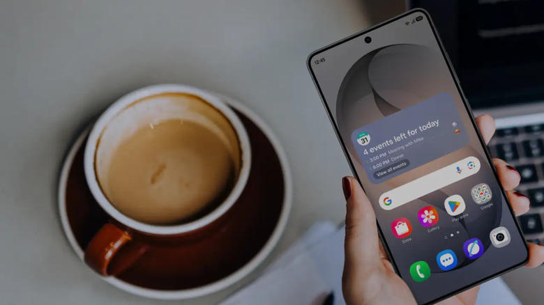 A hand holding a Samsung phone showing the home screen with a cup of coffee and a laptop in the background