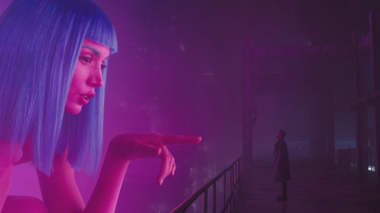 An incredibly tall individual with blue hair stands in front of Ryan Gosling's character K in "Blade Runner 2049"