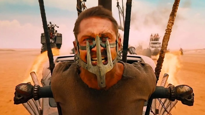 Tom Hardy as Max Rockatansky in "Mad Max: Fury Road," the actor is tied to the front of a vehicle and looks angry about it, they are also wearing a peculiar mask made of metal