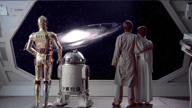 Anthony Daniels as C-3PO, Kenny Baker as R2D2, Mark Hamill as Luke Skywalker, and Carrie Fisher as Princess Leia all stand together aboard a space ship as they gaze at a far away galaxy