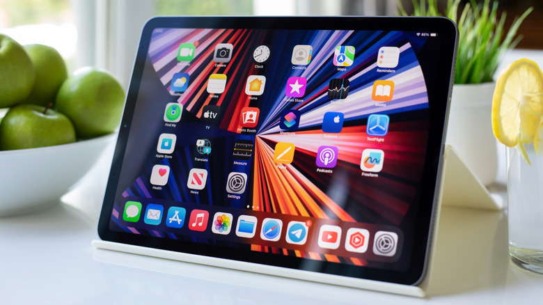 An iPad sitting on a white table between a beverage and a bowl full of green apples, the tablet is displaying the iOS home screen, and there is also a potted plant behind the device