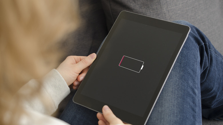 An individual wearing blue jeans and a white sweatshirt has a tablet sitting in their lap, the tablet is displaying a low battery icon