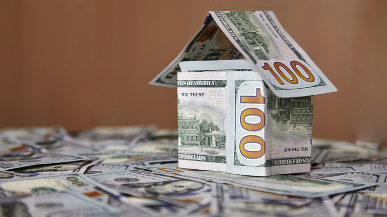 A small house folded from $100 bills sitting on a pile of U.S. currency.