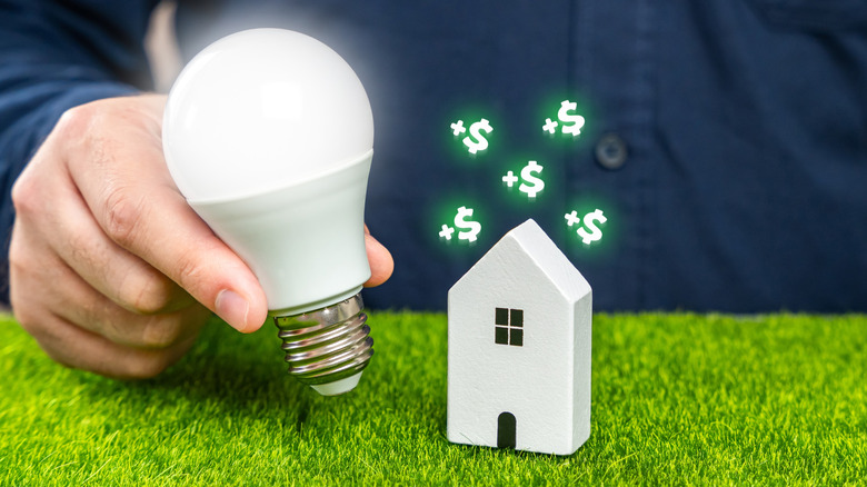 Hand holding a bulb beside a small house, symbolizing energy savings.