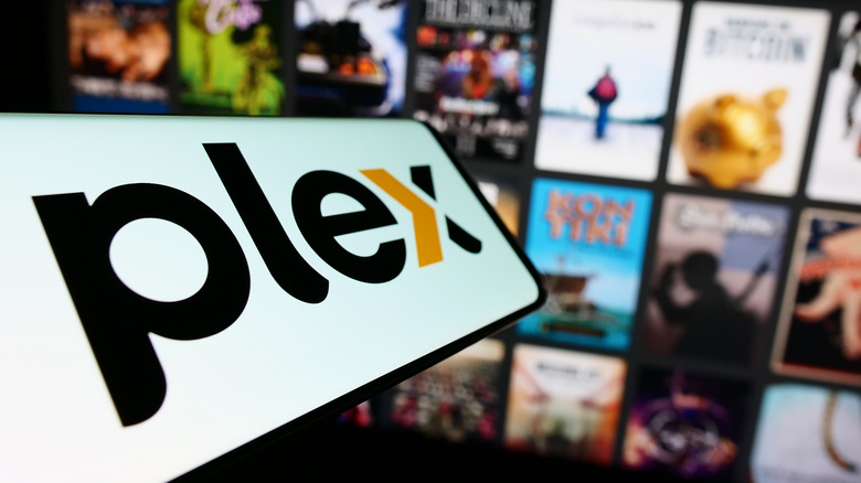 The Plex logo on a smartphone in the foreground. A blurred TV screen showing numerous movies and TV show tiles is the background.