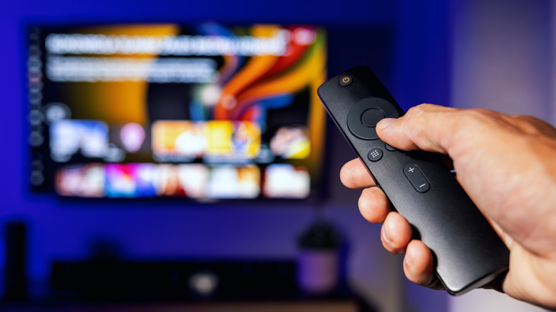 A person pointing a remote toward a blurred TV screen.