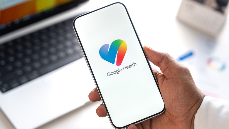 A person's hand by a computer holding a phone showing the Google Health app logo.