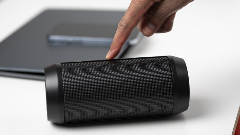 A person turning on a portable speaker on a white table beside a laptop and smartphone