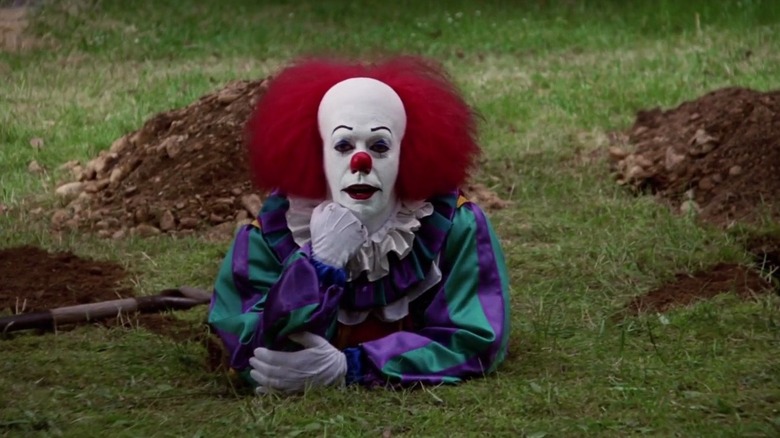 Tim Curry as Pennywise stood in a hole resting his head on his hand in IT