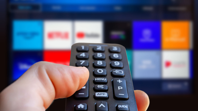 A closeup of a remote control in an individual's left hand, the remote is pointed at a blurry TV screen that is showing multiple icons for familiar streaming services like YouTube and Netflix