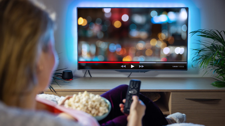 Individual sitting in front of a TV as they hold a remote and a bowl of popcorn, on the TV is a blurred image that appears to be imitating a streaming service