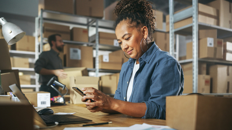 An individuals holds a cellphone in both of their hands as they sit in front of a laptop, the background is filled with many boxes along with an individuals holding a box in their hands