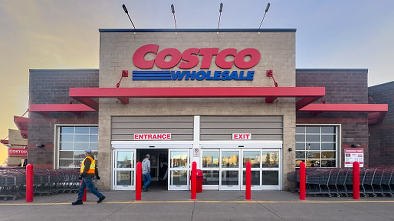 An exterior shot of a Costco Wholesale store, the entire front exterior of the building is prominent, and two individuals as long as two lines of carts can be seen