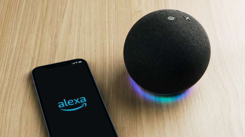 An Amazon Alexa sits on a table next to a smartphone displaying the Alexa logo