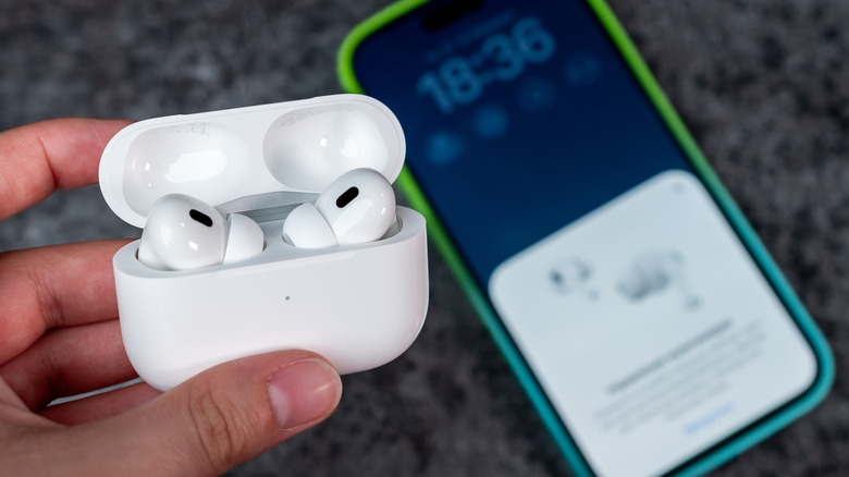 A user's left hand holds the AirPods Pro in their charging case, below the AirPods is an iPhone that is heavily blurred, yet seems to be showing information about the AirPods