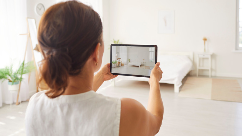 A woman holding a tablet up to take a picture of a white bedroom