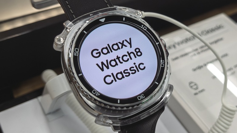 A silver Samsung Galaxy Watch 8 Classic displaying sits on a stand connected to a security device, information about the watch is displayed next to it