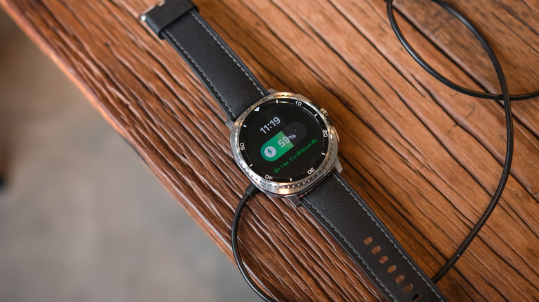 A silver Samsung Galaxy Watch with a black band is connected to a magnetic charging puck as both devices sit on a wooden surface