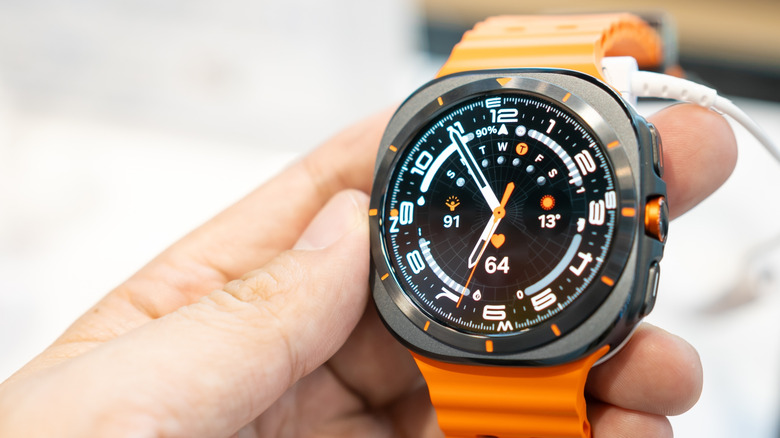 A Samsung Galaxy Watch 7 Ultra being held in a user's right hand, the Watch is connected to either a charging cord of some kind of a security unit